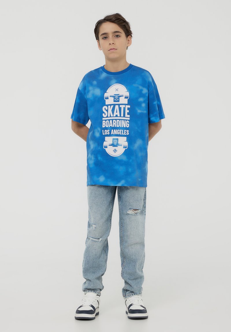 T-shirt blu con motivo tie-dye e grafica bianca "SKATE BOARDING LOS ANGELES", abbinato a jeans chiari strappati e sneakers bianco e nere.