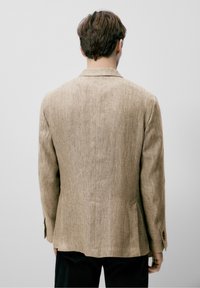 Jasnobrązowa marynarka w jodełkę o dopasowanym kroju, z pojedynczym rozcięciem z tyłu i mankietami na guziki, w zestawie z ciemnymi spodniami.