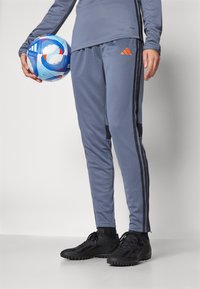 Pantalones deportivos grises con rayas laterales negras, combinados con zapatillas deportivas negras. La persona sostiene un balón de fútbol azul y blanco.