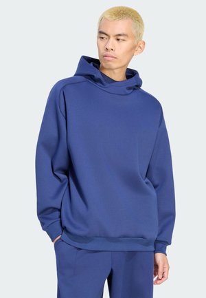 Giovane con capelli biondi corti che indossa una felpa blu larga con cappuccio e pantaloni coordinati, guarda di lato con le mani nelle tasche.
