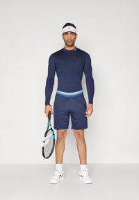 Camisola atlética de manga comprida azul-marinho, calções combinando com uma cintura degradê e uma faixa na cabeça. Segurando uma raquete de ténis, calçando sapatilhas brancas.