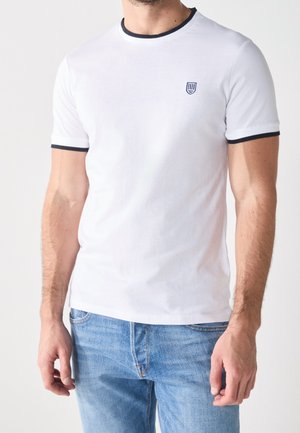 Weißes Baumwoll-T-Shirt mit marinefarbenem Besatz am Kragen und an den Ärmeln. Mit einem kleinen Emblem auf der linken Brust. Kombiniert mit hellblauen Jeans.