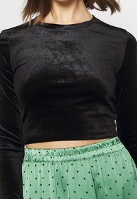 Kvinna i en svart sammet långärmad crop top och en högmidjad grön satinkjol med svarta prickar, visad från halsen till midjan.