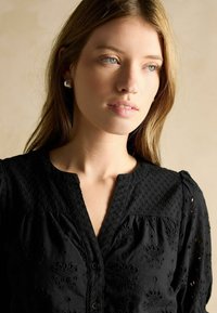 Schwarze Bluse aus strukturiertem Stoff, mit Knopfverschluss und gestickten Blumenmustern. Verfügt über einen runden Halsausschnitt und puffige Ärmel.