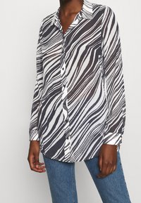 Persona che indossa una camicia a maniche lunghe con stampa zebra, abbinata a jeans blu, in piedi contro uno sfondo chiaro e semplice.