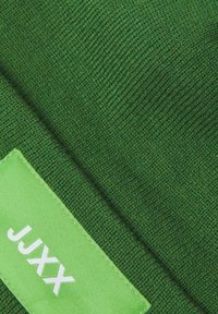 Tessuto lavorato a maglia verde con una consistenza morbida, caratterizzato da un etichetta verde con testo bianco "JXX." Il design complessivo è semplice e utilitario.