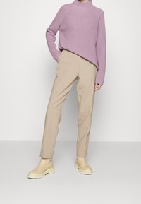 Suéter de punto lila claro con un cuello alto, combinado con pantalones beige de corte ajustado y botas crema sin cordones con suela gruesa.