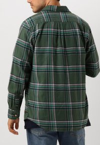 Camisa de cuadros verdes hecha de tela con cuello abotonado, que presenta rayas blancas y azules, y un dobladillo curvado. Se muestra una vista trasera.