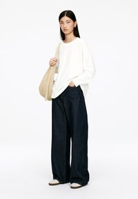 White long-sleeve shirt, dark blue wide-leg jeans, holding a beige shoulder bag, with white sneakers featuring brown soles.
