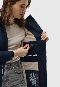 Navy jacket with a soft beige lining, featuring a pocket with a red pull tab, logo accents, and distinctive quilted details on the sleeve.
