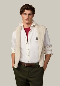 Suéter de punto crema usado sobre una camisa blanca de botones y una camiseta roja, combinado con pantalones verde oscuro y cinturón marrón, sobre un fondo neutral.