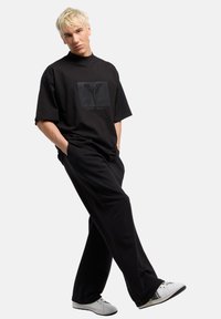 Juodas oversized t-shirt su tekstūruotu logotipo spaudiniu, derinamas su laisvomis juodomis kelnių ir baltais batais. Minimalistinis, laisvas dizainas.