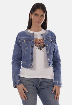 Donna con capelli lunghi e scuri che indossa una giacca di jeans blu con strass, una maglietta bianca con un motivo a cuore multicolore e jeans blu.