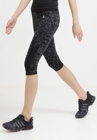 Persona che indossa leggings neri con stampa leopardata a tre quarti e scarpe da corsa nere mentre cammina su uno sfondo bianco uniforme.
