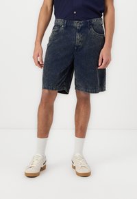 Pantalones cortos de mezclilla en azul oscuro con una sutil textura desgastada, cuentan con bolsillos y una cinturilla estándar. Se llevan con calcetines claros y zapatillas casuales.