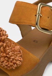 Πορτοκαλί πλατφόρμα πέδιλο από σουέντ με φαρδιά λουρίδα που φέρει χρυσή αγκράφα και διακοσμητικά με άνθη πομ πομ στο μπροστινό μέρος. Η σόλα είναι σε ταμπά χρώμα.