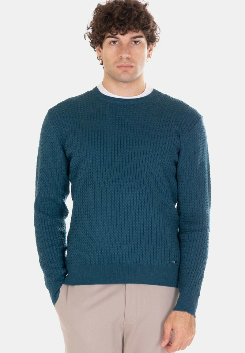 Maglione lavorato a maglia verde petrolio con motivo strutturato e scollo rotondo, caratterizzato da polsini e orlo a coste. Abbinato a pantaloni di colore chiaro.