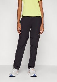 Schwarze Zip-off-Hose aus leichtem Stoff, mit schmal zulaufender Form, Seitentaschen und einem kleinen Logoakzent. Kombiniert mit grauen Sportschuhen.