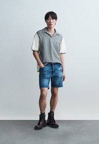 Fiatal férfi áll egy sima fal előtt, két árnyalatú rövid ujjú inget, farmer rövidnadrágot, fekete zoknit és sötétbarna fűzős bakancsot viselve.