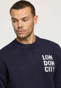 Pull en tricot bleu marine avec un col ras du cou côtelé et un graphique en texte blanc "LONDON CITY" sur la poitrine gauche. Texture douce et coupe ajustée.