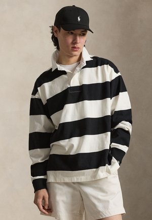 Ung person som har på seg en svart og hvit bredstripet rugbytrøye, beige shorts og en svart baseballcaps med en liten hvit logo.