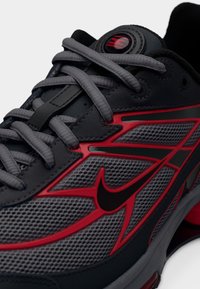 Αθλητικό παπούτσι Nike με γκρι πλέγμα στο πάνω μέρος, κόκκινες λεπτομέρειες, υφασμάτινες επενδύσεις με υφή, μαύρο λογότυπο και επίπεδα γκρι κορδόνια. Σχεδιασμός με ελαφρώς στρογγυλεμένη μύτη.