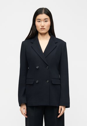 Femeie cu păr lung și negru, purtând un blazer negru cu două rânduri de nasturi, având patru nasturi și buzunare cu clapetă, stând în picioare în fața unui fundal simplu.