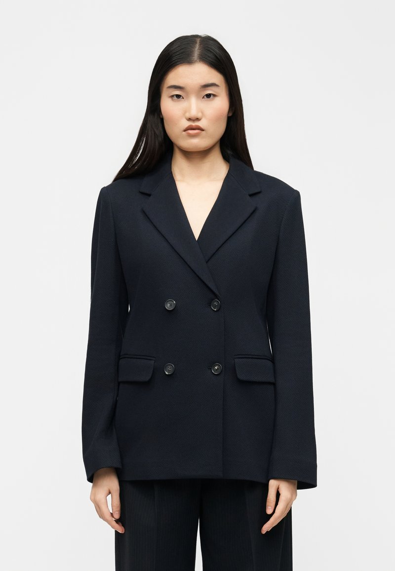 Γυναίκα με μακριά μαύρα μαλλιά που φοράει μαύρο σακάκι με διπλή σειρά κουμπιών, τέσσερα κουμπιά και τσέπες με καπάκι, στέκεται μπροστά σε απλό φόντο.