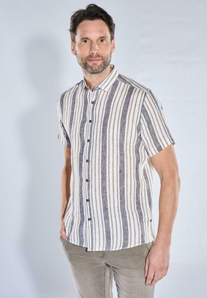 Mann mit kurzen dunklen Haaren und Bart, der ein kurzärmeliges Hemd mit vertikalen grauen, weißen und beigen Streifen trägt und mit einer Hand in der Tasche steht.