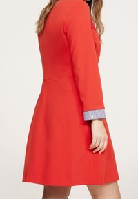 Rotes Kleid mit langen Ärmeln, tailliertem Oberteil, schwingendem Rock und einem kontrastierenden gestreiften Bund. Hergestellt aus einem glatten, strukturierten Stoff.