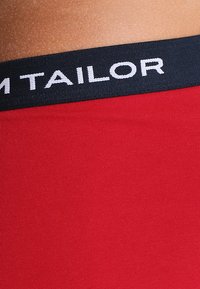 Roter Stoff mit glatter Textur, ausgestattet mit einem schwarzen elastischen Bund, der in weißer Schrift mit "M TAILOR" beschriftet ist.