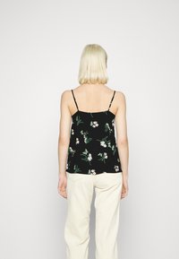 Top senza maniche nero con motivo floreale, caratterizzato da spalline sottili, una vestibilità ampia e un design di foglie verdi e fiori rosa chiaro. Indossato con pantaloni beige.
