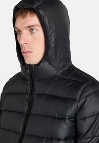 Närbild av en ung man som bär en svart dunjacka med huva. Hans uttryck är neutralt, och han blickar något åt sidan.