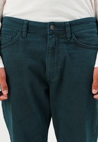 Homme portant un jean en denim bleu foncé avec des poches avant et une chemise blanche à manches longues, les mains détendues sur les côtés.
