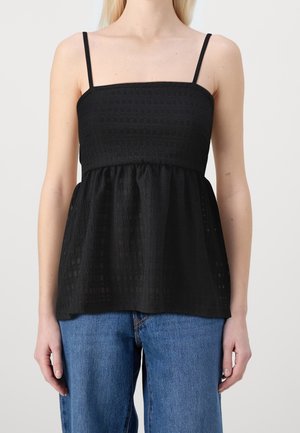 Femme portant un haut sans manches texturé noir avec de fines bretelles et un jean bleu, montrée du cou aux hanches sur un fond uni.