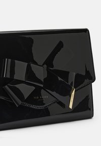 Pochette en cuir verni noir avec un grand nœud. Présente des éléments en or avec le logo "TED BAKER". Texture lisse et forme structurée.