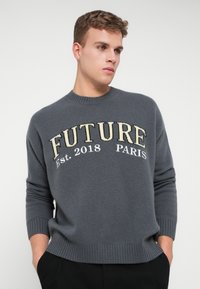 Giovane uomo con un maglione grigio con la scritta "FUTURE Est. 2018 PARIGI", in piedi con le mani in tasca, che guarda di lato su uno sfondo bianco.