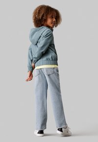 Lyseblå letvægtsjakke med elastiske manchetter og kant, parret med en gul t-shirt og lyse vaskede straight-leg jeans. Hvide sneakers.