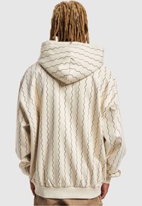 Beige hettegenser med bølgete svart linjemønster, med en romslig hette, ribbede mansjetter og nederkant, laget av mykt stoff for komfort.