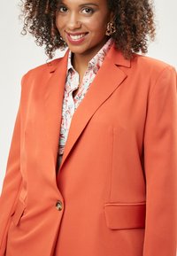 Orange Blazer mit einem einzelnen Knopf, mit Reverskragen und zwei vorderen Taschen, kombiniert mit einem floral gemusterten Hemd darunter.