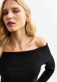 Blond kvinna med guldörhänge och en svart ribbad off-shoulder-tröja, ser något åt vänster mot en enkel vit bakgrund.