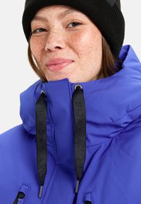 Blaue isolierte Jacke mit hohem Kragen, schwarzen Logoakzenten und verstellbaren Kordelzügen. Sie zeichnet sich durch eine glatte Textur und ein elegantes Design aus.