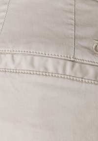 Tissu beige à texture lisse, présentant une couture froncée et des coutures délicates. Comprend des détails brodés subtils.