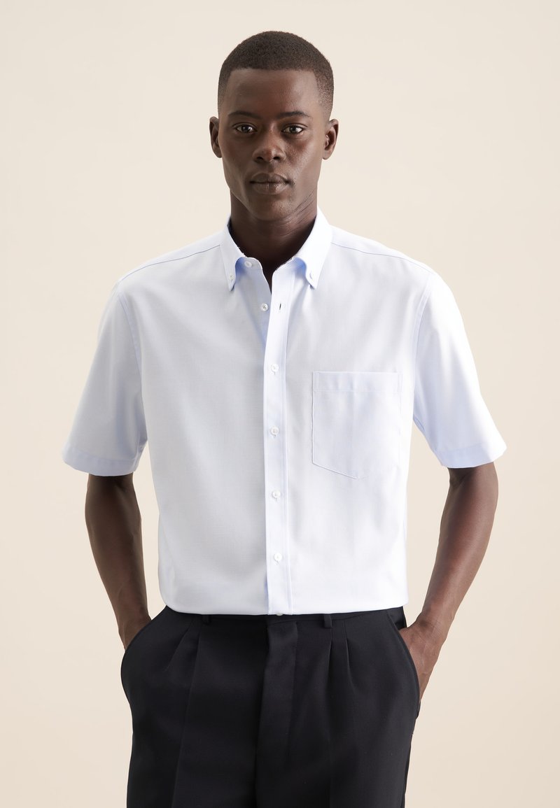 Camicetta azzurra chiara a maniche corte con colletto alla francese, dotata di taschino sul lato sinistro e una texture liscia e intrecciata. Abbinata a pantaloni scuri.