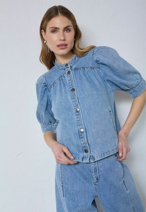 Pikkade juustega naine, kes kannab pärlikeesid, hele sinist denimist nööbitavat särki pahtlike varrukatega ja sobivaid denimpükse ühevärvilise taustaga.