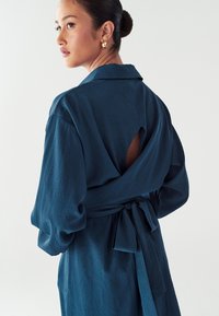 Temnoč teal obleka z dolgimi rokavi, teksturirana tkanina, izrez na hrbtu in zavezan pas. Oblikovan ovratnik poudarja obliko.