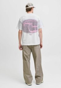 Hvid kortærmet t-shirt med et pink grafisk tryk af træer på ryggen; parret med beigefarvede vide bukser og en lyse farvet kasket.
