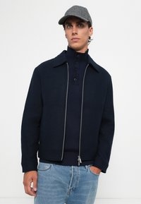 Young man wearing a gray cap, navy zip-up jacket over a navy ribbed sweater with buttons, and light blue jeans, standing against a white background.