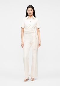 Mujer con cabello largo y oscuro, vestida con un mono de manga corta color crema con cinturón lateral y tacones abiertos beige, de pie frente a un fondo liso.