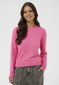 Femme souriante avec de longs cheveux noirs, portant un pull rose texturé et une jupe en denim gris délavé, se tenant devant un fond uni.